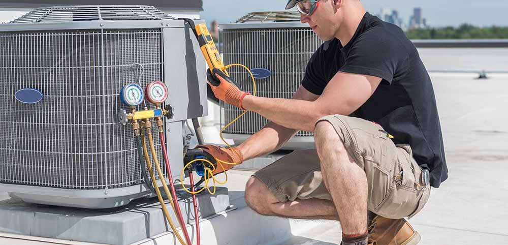 A professional HVAC technician inspecting ductwork and checking measurements on a digital tablet next to an outdoor AC unit, with sunlight in the background and tools neatly arranged nearby.