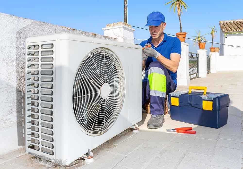 A close-up of an outdoor AC unit with a technician inspecting it, capturing a faint mist escaping from a pipe, with clear skies in the background and subtle toolkits beside the unit.