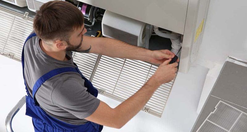 A technician repairing a central AC unit, with tools and diagnostic equipment visible.