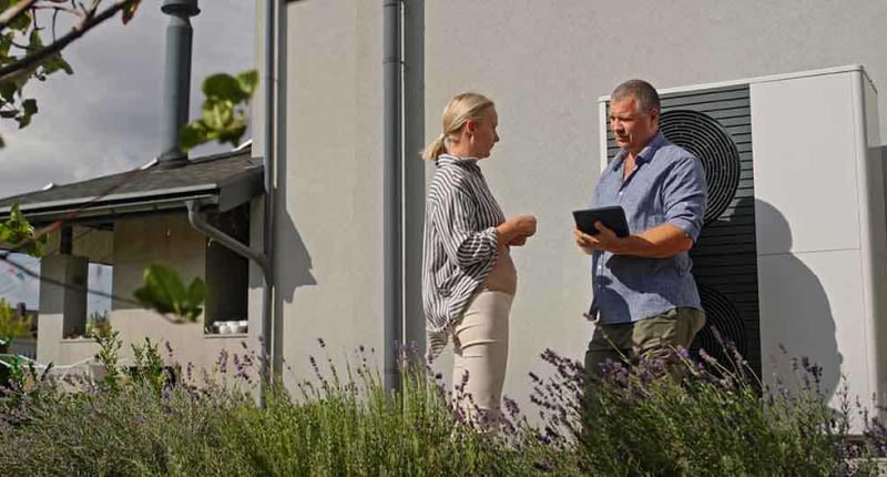 A technician discussing documents with a homeowner. In the background, a new heat pump installation can be seen.